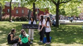Trois élèves tiennent une pancarte sur laquelle il est écrit "Casse-tête des récompenses", et présentent leur projet à deux femmes assises dans un parc.
