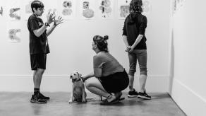 Un élève discute avec une femme qui flatte un chien. Une autre femme regarde les dessins exposés.