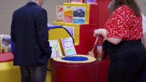 Deux personnes regardent des livres crées en collaboration avec les élèves, présenté sur du matériel de gymnase coloré. Crédit photo @ Éloi Angers-Roy.