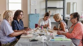Les artistes assises autour d'une table en compagnie des participantes aînées, pour l'atelier de céramique. Crédit photo : Katya Konioukhova.